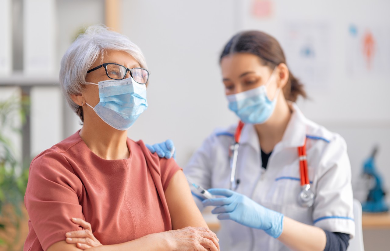 Eine mittlelalte Frau mit Brille trägt eine OP-Maske und wird von einer Ärztin geimpft